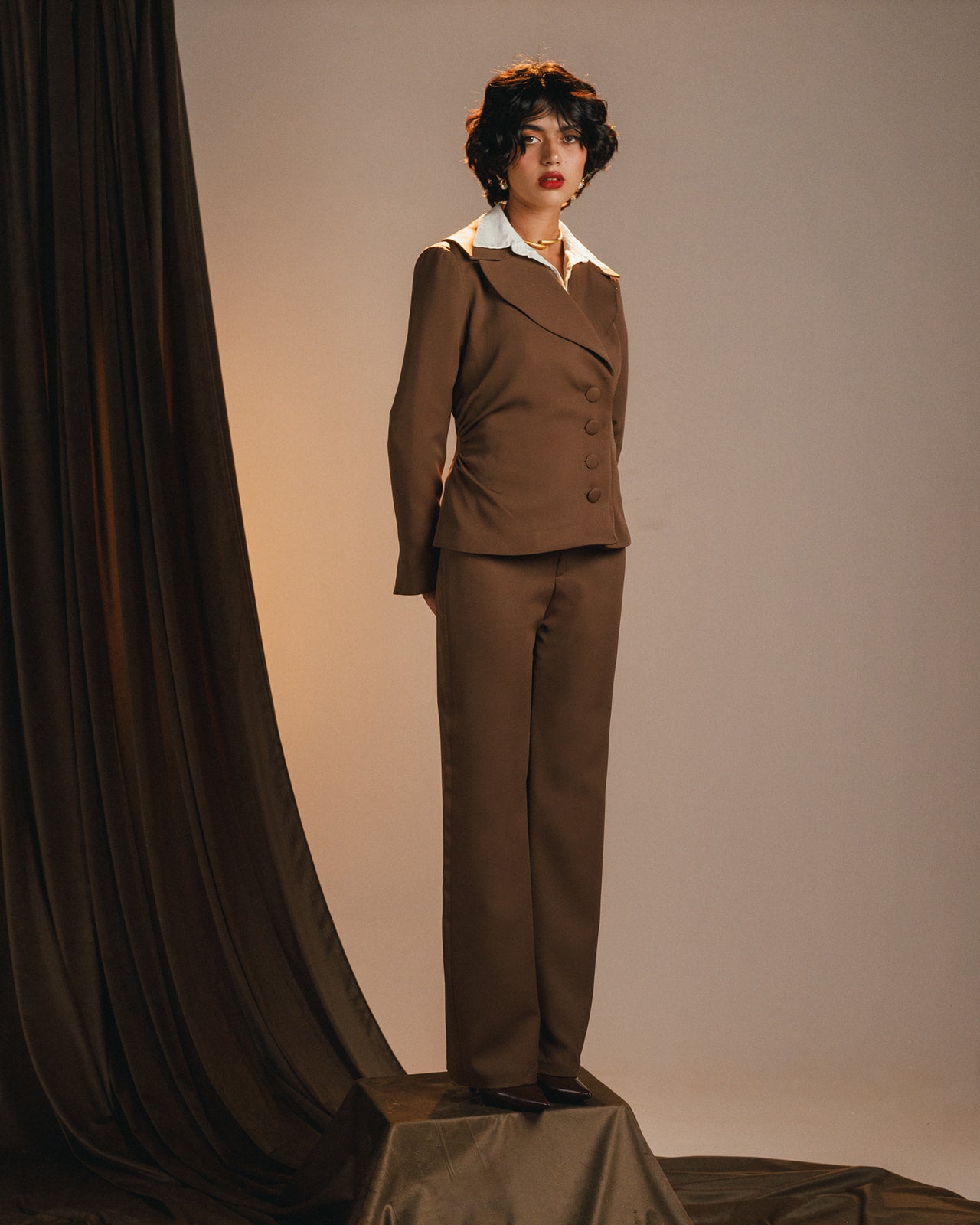 Woman wearing a brown suit with a white shirt and red lipstick against a plain background